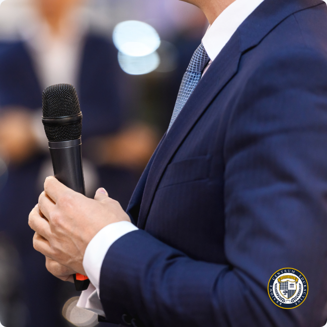 Zbliżenie na mężczyznę w eleganckim, granatowym garniturze z białą koszulą i krawatem w drobny wzór, trzymającego w prawej dłoni czarny mikrofon. Kadrowanie skupia się na jego torsie i ręce, sugerując moment wystąpienia publicznego.