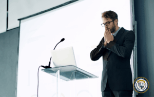 Mężczyzna w garniturze i okularach przy mównicy z laptopem, sprawia wrażenie zdenerwowanego lub skupionego (trema) przed prezentacją.