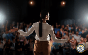 Kobieta stoi na scenie tyłem do widza, gestykulując rękami do dużej, rozmytej publiczności oświetlonej reflektorami.