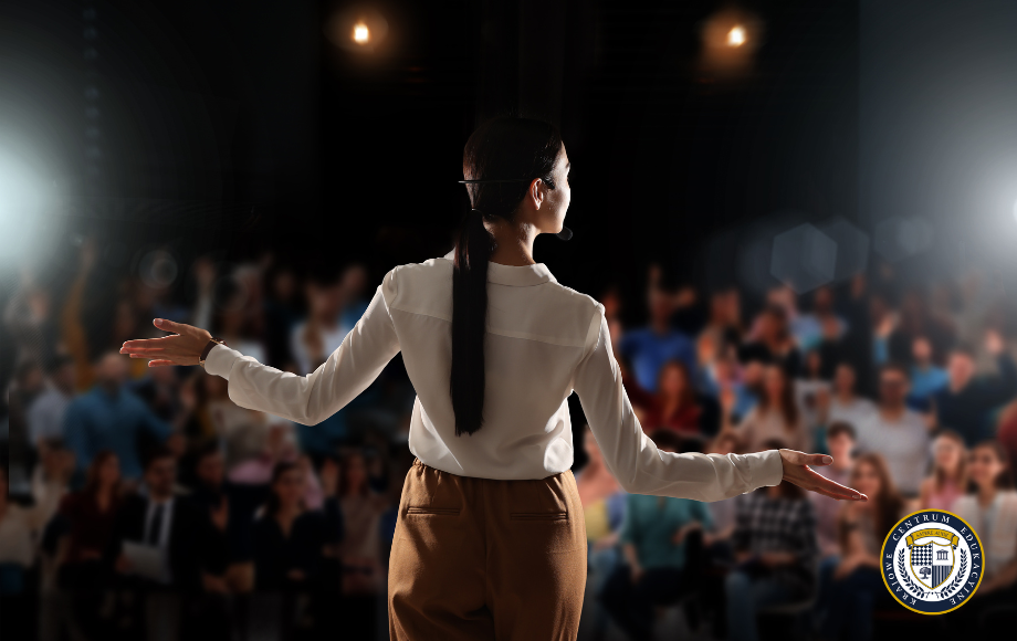 Kobieta stoi na scenie tyłem do widza, gestykulując rękami do dużej, rozmytej publiczności oświetlonej reflektorami.