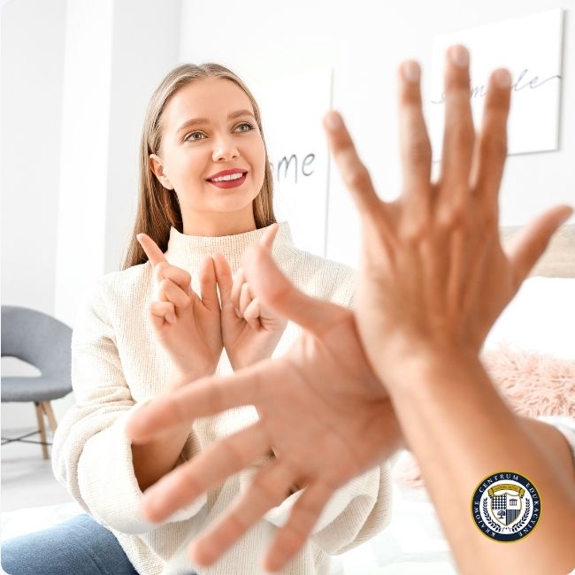 Uśmiechnięta kobieta w beżowym swetrze demonstruje mowę rąk, pokazując gesty dłońmi w jasnym wnętrzu.
