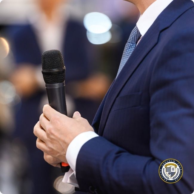 Mężczyzna w granatowym garniturze trzymający mikrofon podczas wystąpienia publicznego, w tle rozmyte sylwetki publiczności.