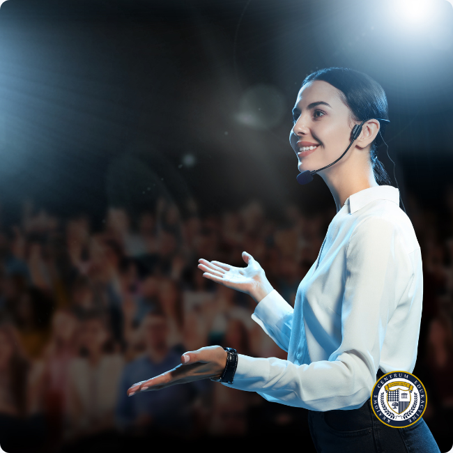 Kobieta w białej koszuli i z mikrofonem nagłownym stoi na scenie, patrząc w lewo i uśmiechając się. Używa otwartej, dynamicznej gestykulacji obiema dłońmi, angażując w ten sposób publiczność.