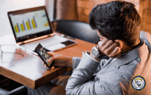 Mężczyzna w garniturze prokrastynuje, siedząc przy laptopie z wykresami, lecz rozprasza się, intensywnie używając smartfona.