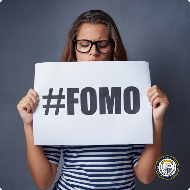 obieta w okularach i koszulce w paski trzyma przed sobą kartkę z napisem #FOMO.