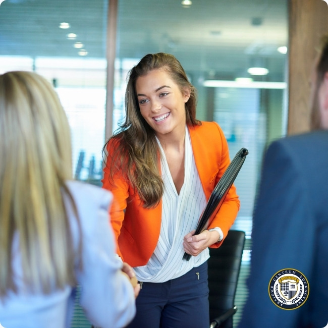 ALT:Uśmiechnięta kobieta w pomarańczowej marynarce wita się i robi dobre pierwsze wrażenie podczas spotkania biznesowego w biurze