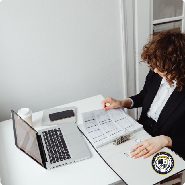 Kobieta w garniturze siedząca przy biurku analizuje dokumenty i wykresy obok laptopa, ilustrując proces rozliczania i ewidencjonowania godzin nadliczbowych