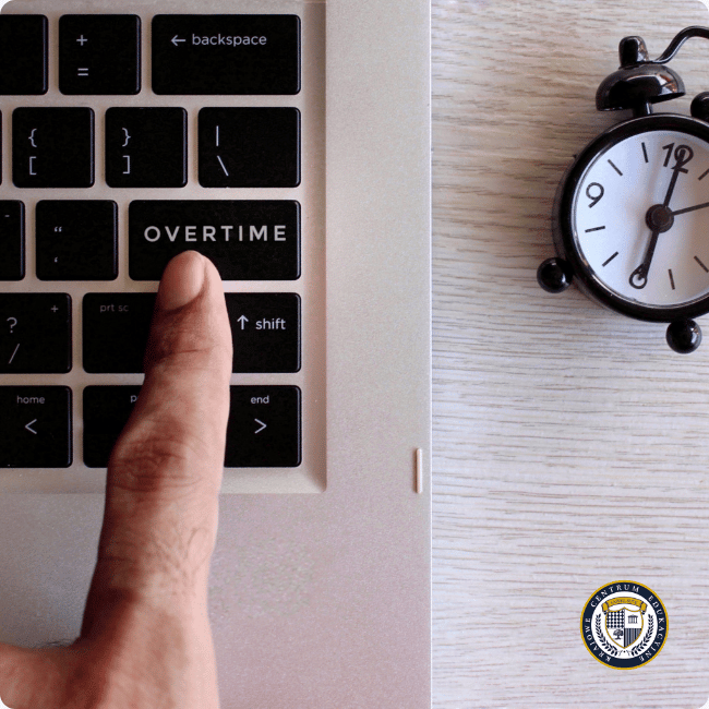 Palec wskazujący naciskający klawisz "OVERTIME" na klawiaturze laptopa, obok budzik pokazujący godzinę 12